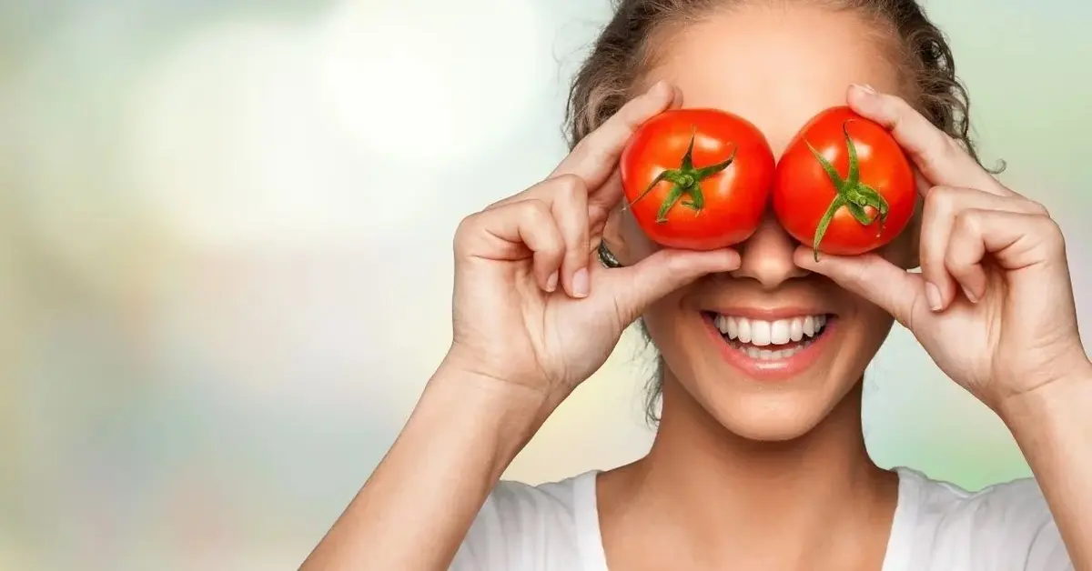 Kobieta, radośnie i szeroko uśmiechnięta, zasłania oczy pięknymi czerwonymi pomidorami