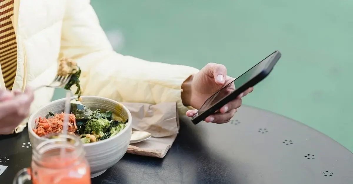 Sylwetka człowieka siedzącego przy czarnym stoliku, przeglądającego telefon przy misce z warzywami i szklance soku