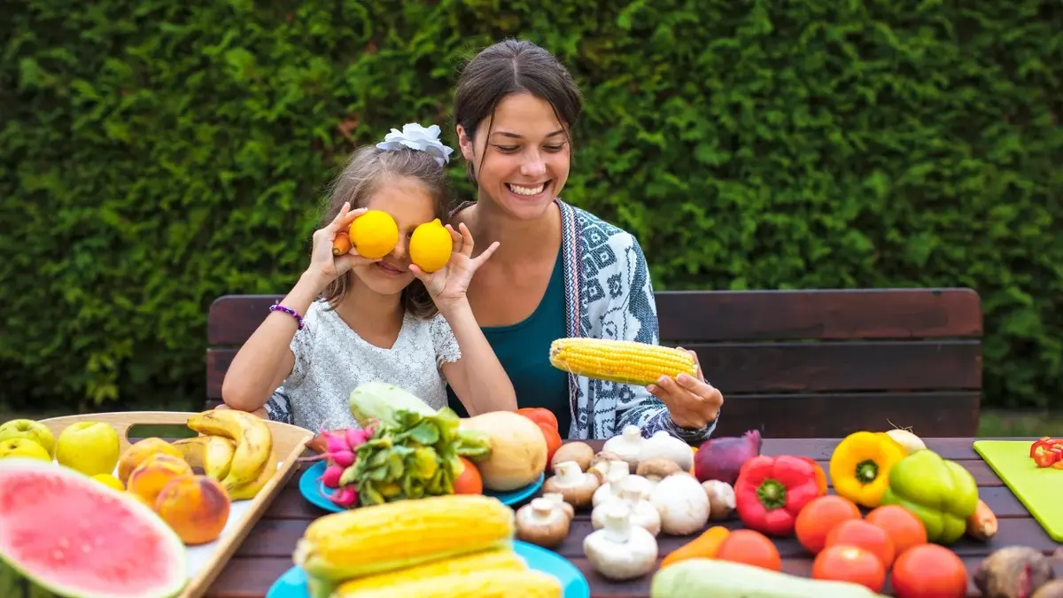 owoce i warzywa na stole z tyłu kobieta z dzieckiem które trzyma przy oczach pomarańcze za nimi tło z roślin