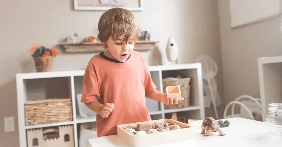 Mały chłopiec z blond włoskami i łososiowej bluzie bawi się radośnie stojąc w pokoju dziecięcym przy białym stoliku