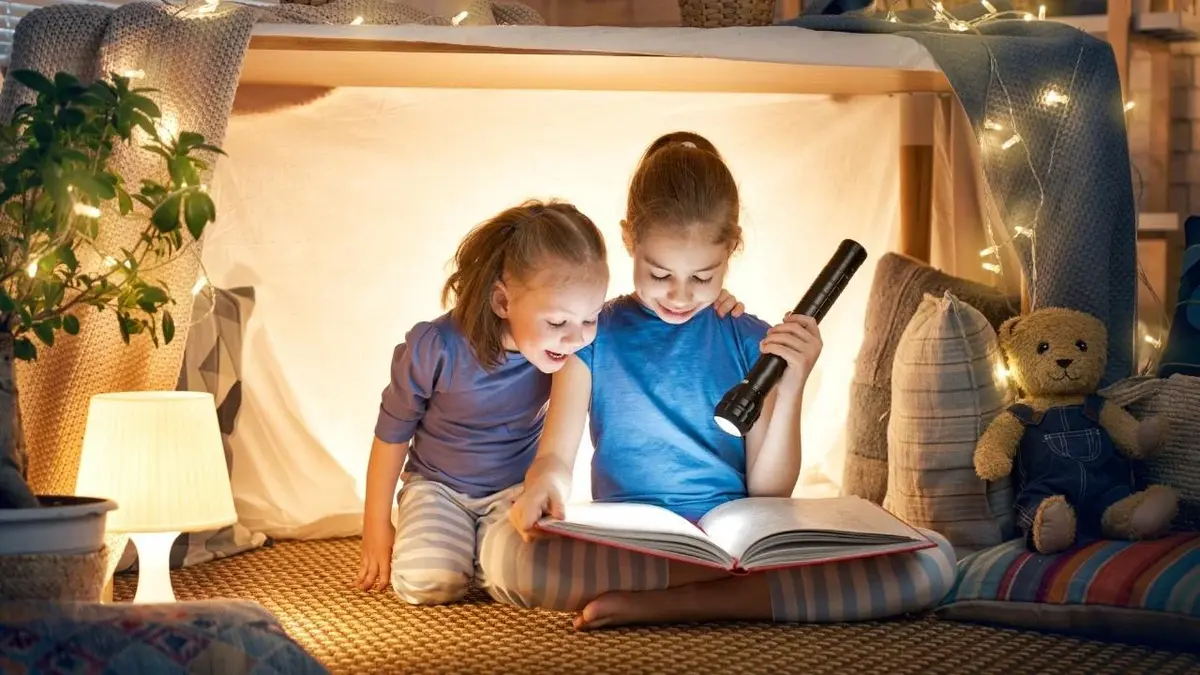 Dwie dziewczynki czytają książkę oświetlając ją latarką.