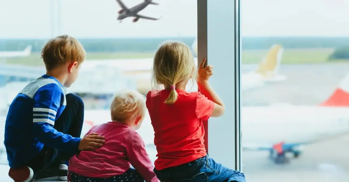 Troje dzieci wygląda przez szybę na lotnisku i obserwuje lecący samolot.
