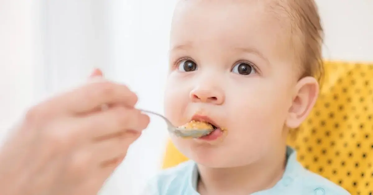 Małe dziecko w trakcie rozszerzania diety, jest karmione łyżeczką. Ma niepewną minę.