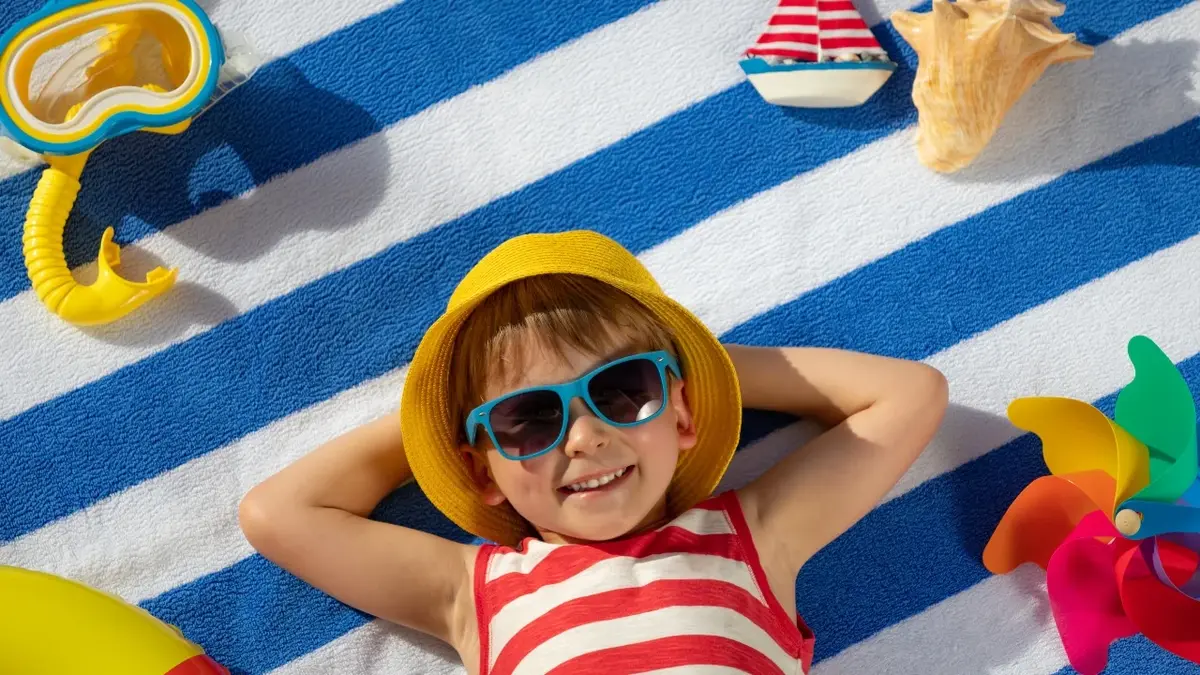 Chłopiec w kostiumie kąpielowym, kapeluszu i okularach przeciwsłonecznych leży na kocu.