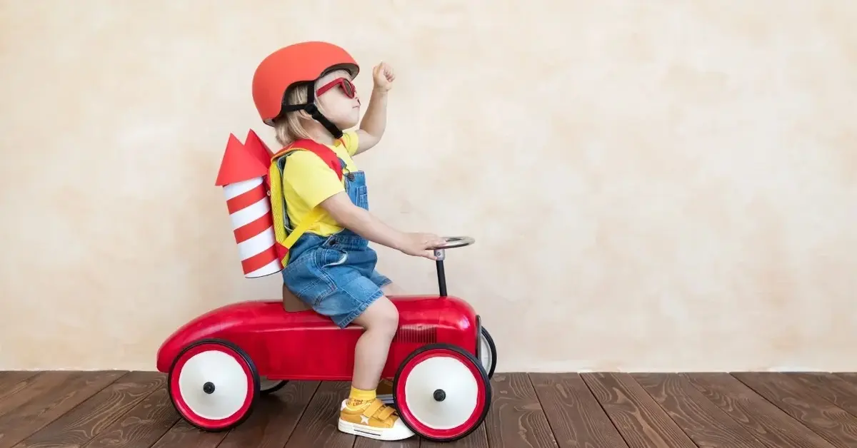 Dziecko w kasku jeździ na zabawkowym pojeździe. Dziecko ma na przyczepione na plecach papierowe rakiety  kosmiczne.