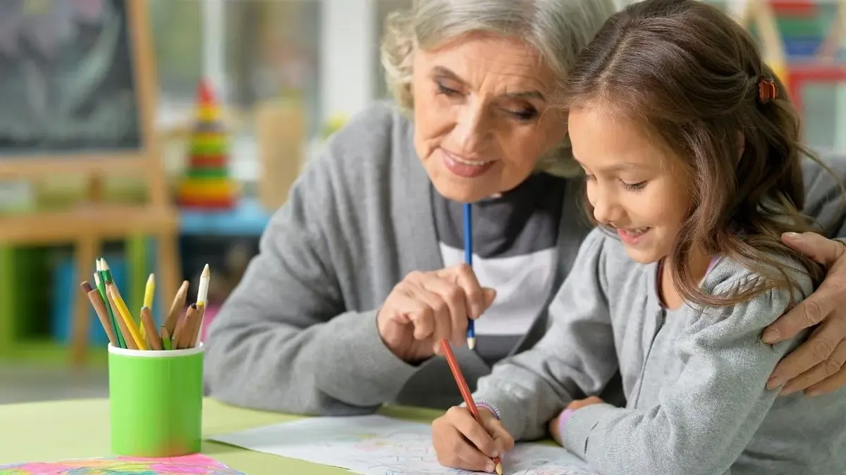 Dziecko siedzi w domu przy stoliku i rysuje razem z babcią.