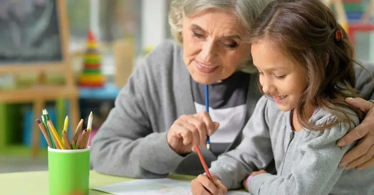 Dziecko siedzi w domu przy stoliku i rysuje razem z babcią.
