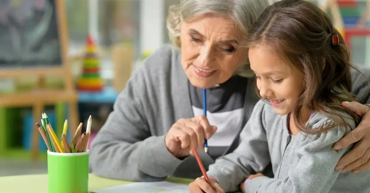 Dziecko siedzi w domu przy stoliku i rysuje razem z babcią.
