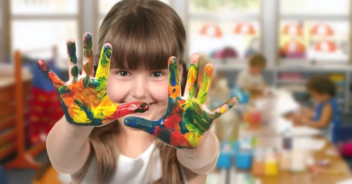 Uśmiechnięta dziewczynka w przedszkolu ma dłonie pobrudzone farbami.