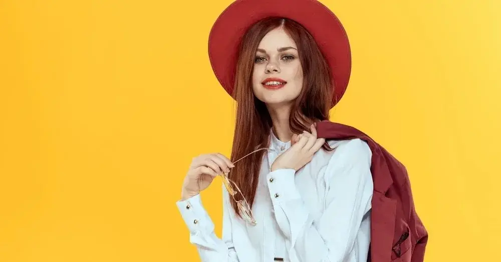 Na intensywnie żółtym tle kobieta w czerwonym kapeluszu z zarzuconym na ramię żakietem w białej koszuli na wymiar