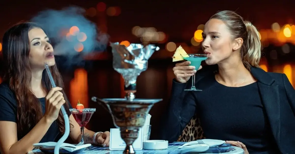 Dwie kobiety ubrane imprezowo w knajpie popijają kolorowe drinki oraz palą fajkę wodną