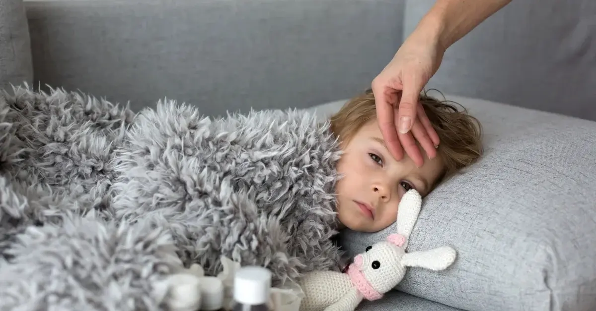Dziecko przykryte kocem leżące w łóżku, którego czoła dotyka ręka sprawdzając temperaturę. Koło dziecka leży pluszowy kr