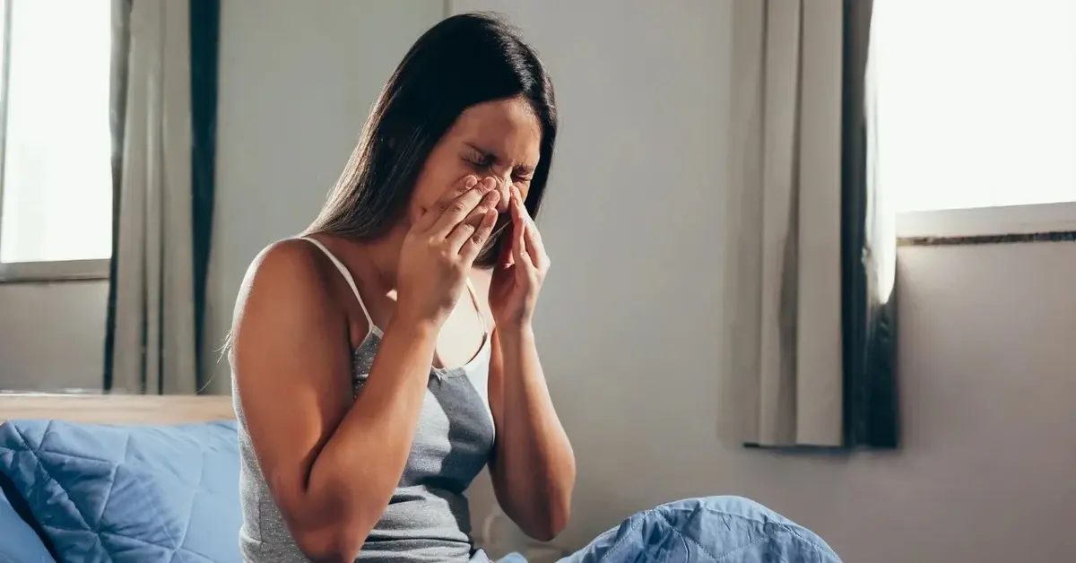 Kobieta siedząca na łóżku i trzymająca dłonie na twarz w okolicach nosa 
