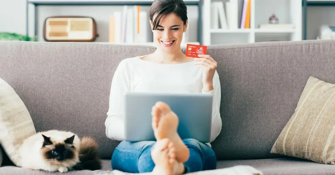 Na kanapie w salonie siedzi uśmiechnięta kobieta robiąc zakupy online przy użyciu cashback i oszczędzając