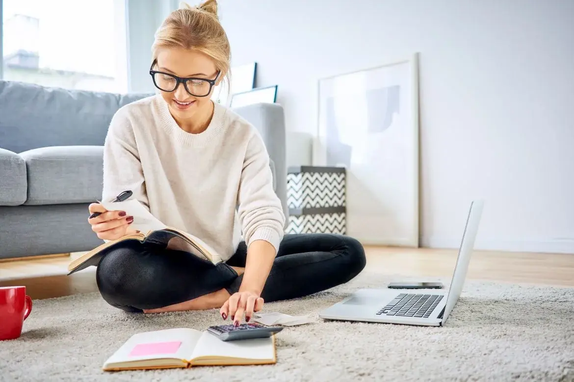 Na dywanie siedzi uśmiechnięta kobieta w okularach robiąc zakupy online przy użyciu cashback