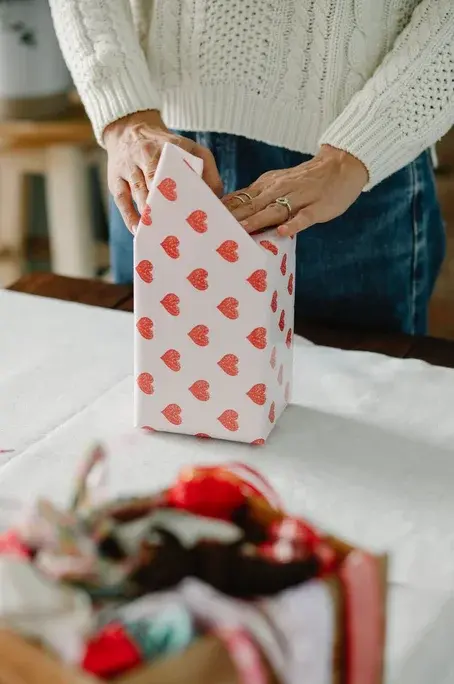 Przy białym stole kobiece dłonie kończą pakować prezent w biały papier w czerwone serduszka