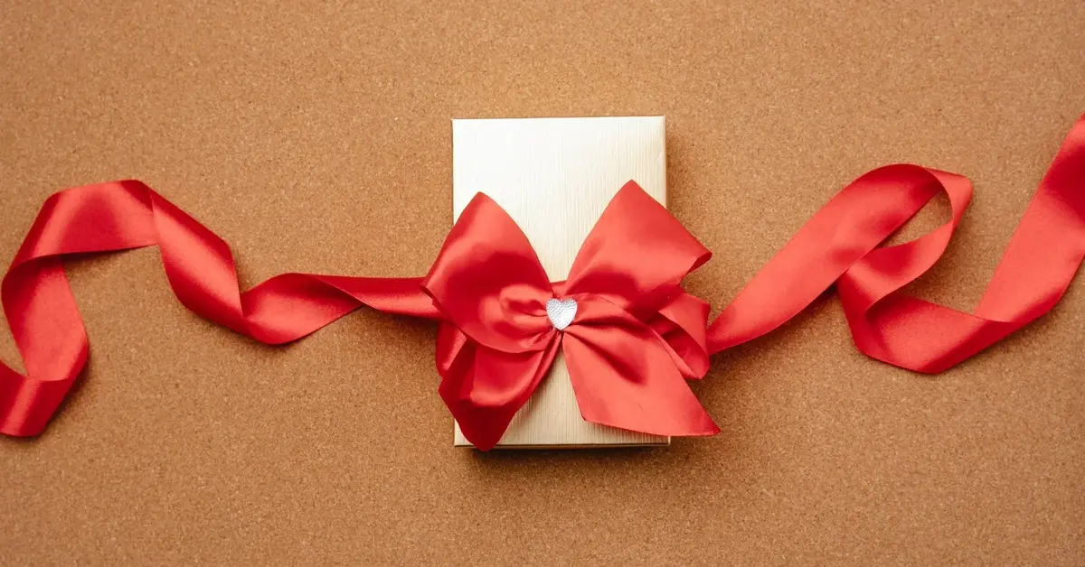 Na neutralnym tle prezent zapakowany w biały papier z fantazyjnie przewiązaną czerwoną wstążką