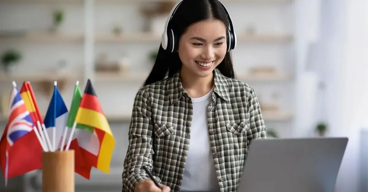 kobieta ze słuchawkami uśmiecha się do laptopa obok ma flagi różnych państw