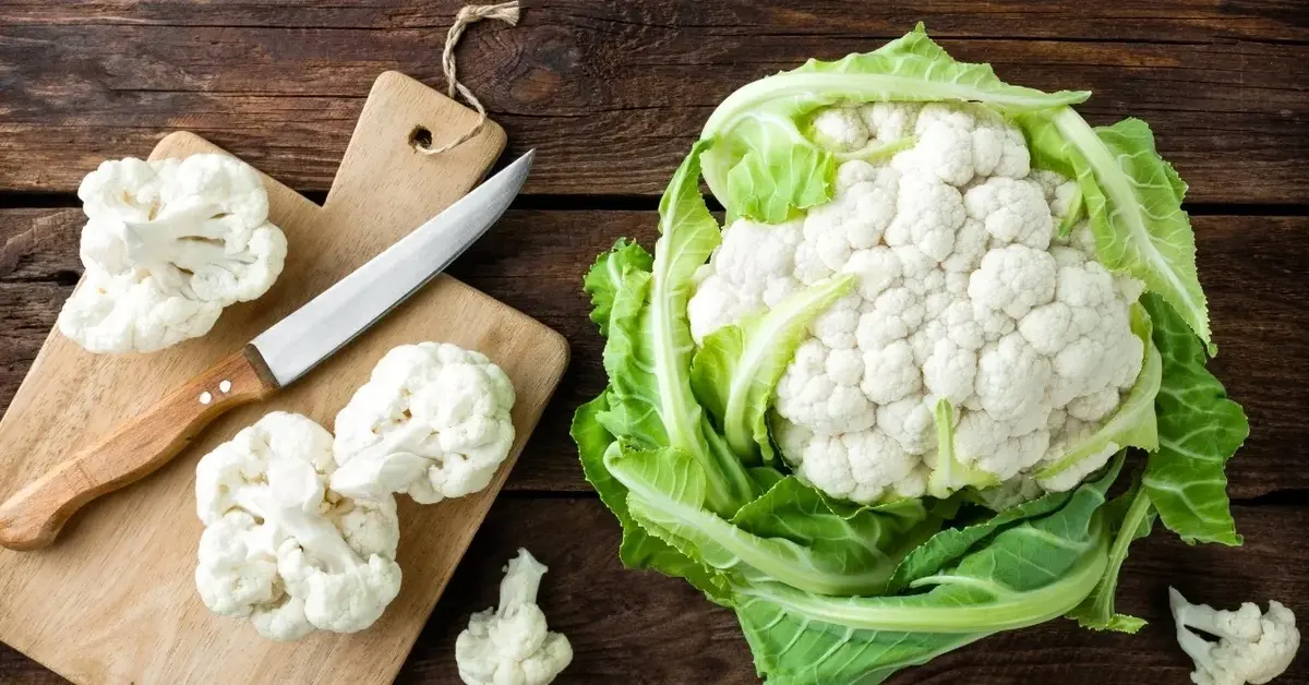 Kalafior leży na drewnianym blacie. Obok deska, różyczki kalafiora i nóż.