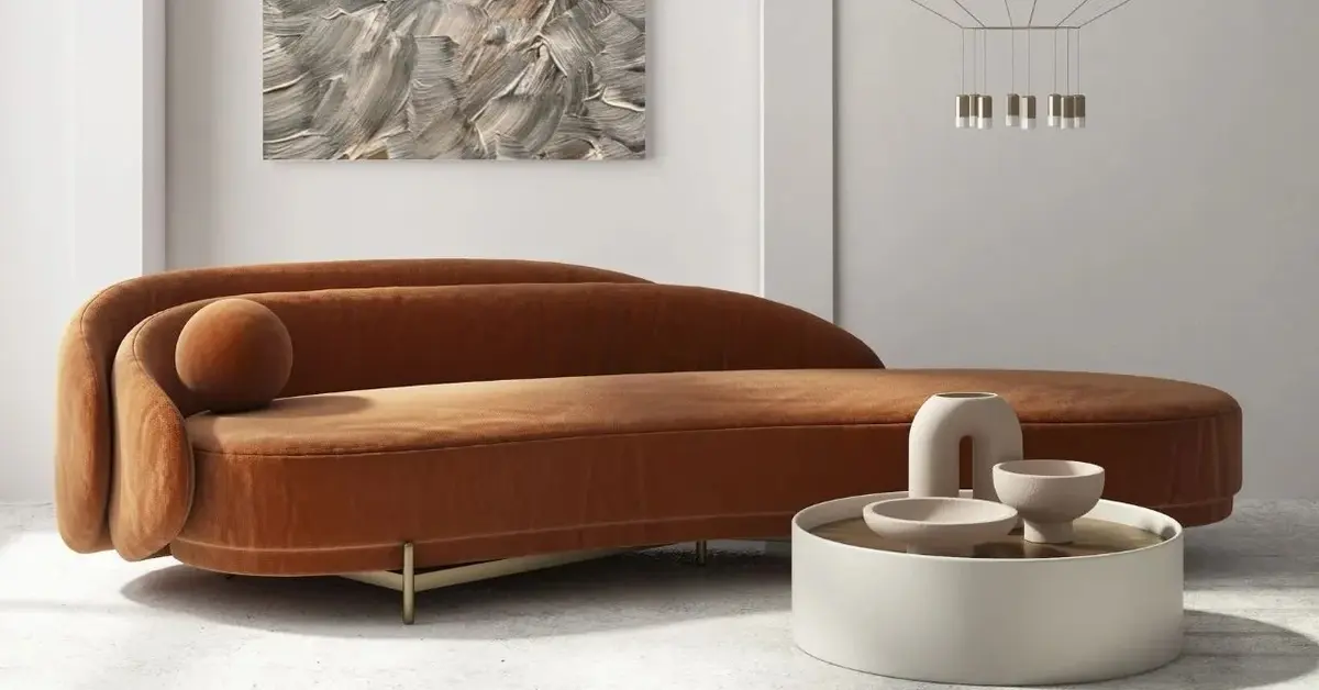 Wygięta karmelowa kanapa z podwójnym oparciem jako statement piece w białym salonie koło białego stolika z wazonami