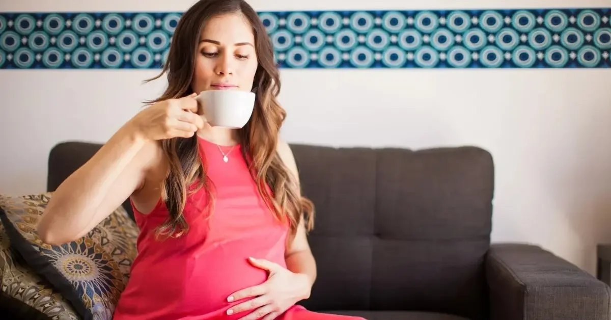 kobieta w ciąży w czerwonej sukience trzyma sie za brzuch w drugiej ręce ma kubek z kawą