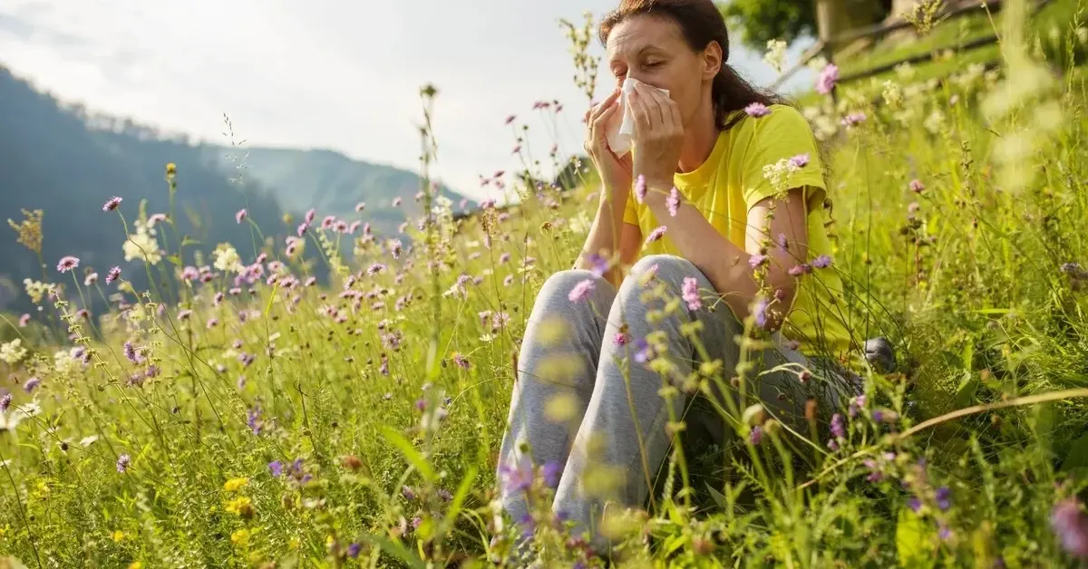 Kobieta siedzi na łące pełnej kwiatów i dmucha nos z powodu alergii.