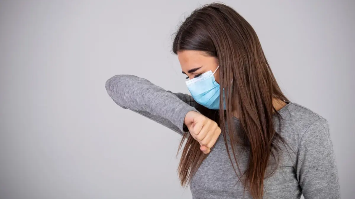 Kobieta z zapalniem płuc w maseczce kaszle, zasłaniając łokciem twarz.