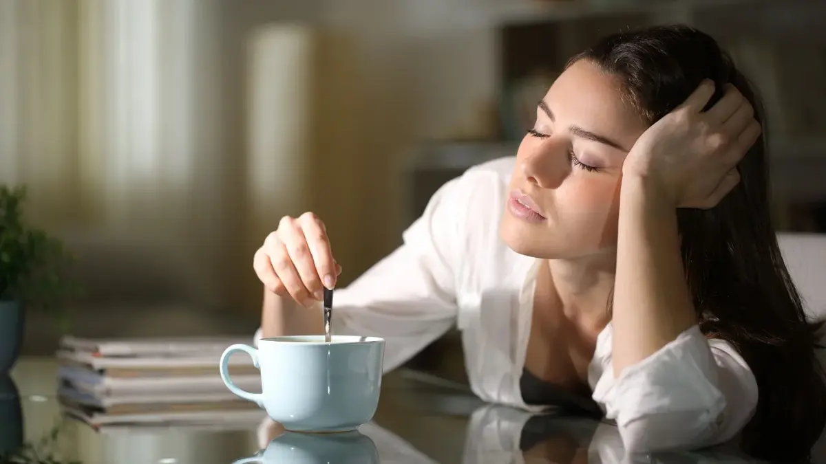 Zaspana kobieta z zamkniętymi oczami, miesza kawę w filiżance.