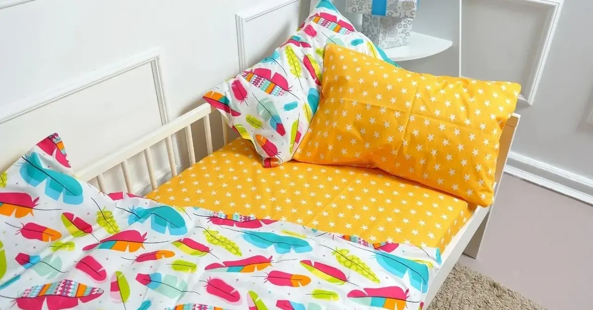 W białym dziecięcym pokoiku, na białym łóżeczku żółte prześcieradło i żółta poduszka oraz pościel w kolorowe piórka