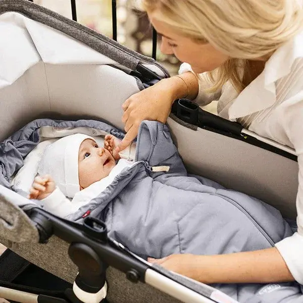 Mama długie blond włosy z czułością spogląda na noworodka leżącego w wózku z wyprawki
