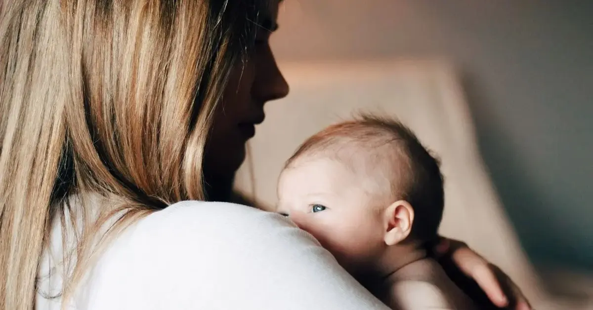 Mama o długich blond włosach w jasnej bluzce przytula do ramienia słodkiego noworodka