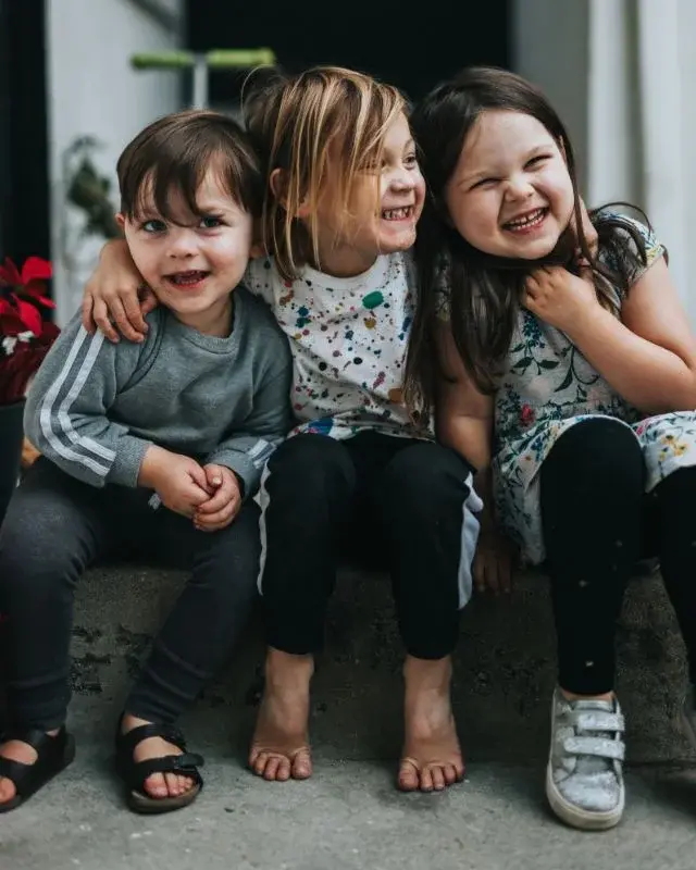 Troje dzieci radośnie uśmiechniętych w wieku przedszkolnym siedzi na stopniu obejmując się