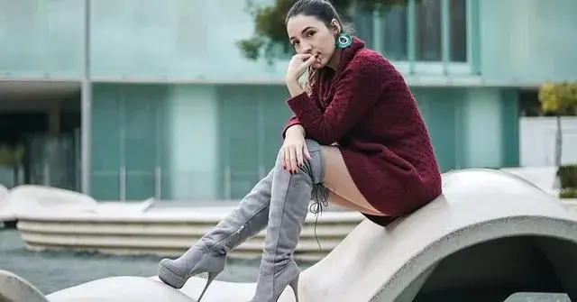 Na falstej awangardowej łąwce siedzi kobieta w bordowym swetrze i wysokich za kolona popielatych kozakach zamszowe