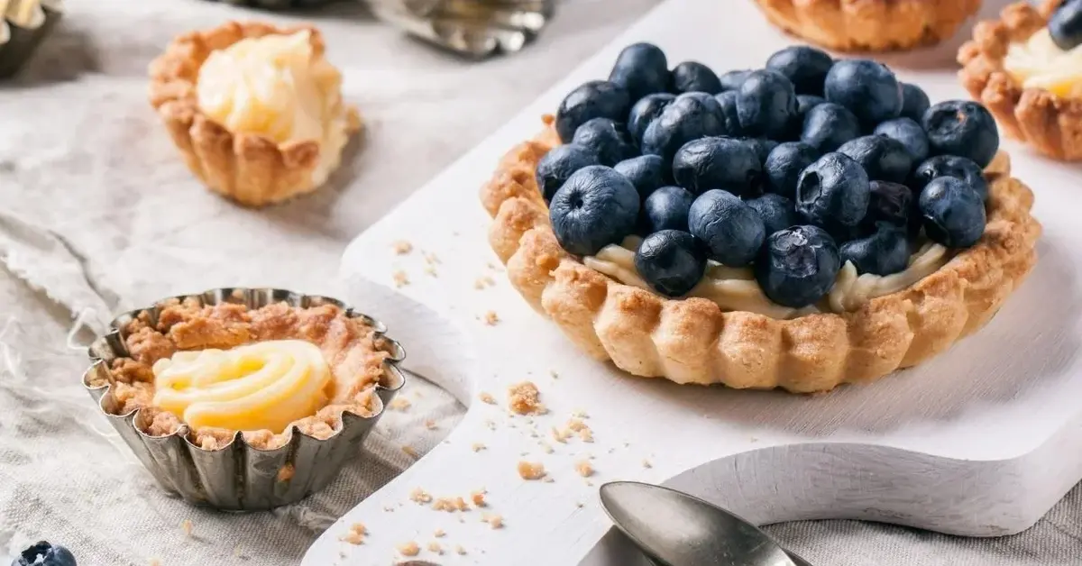 Krucha babeczka z borówkami i kremem waniliowym na białej desce. Obok babeczka w foremce