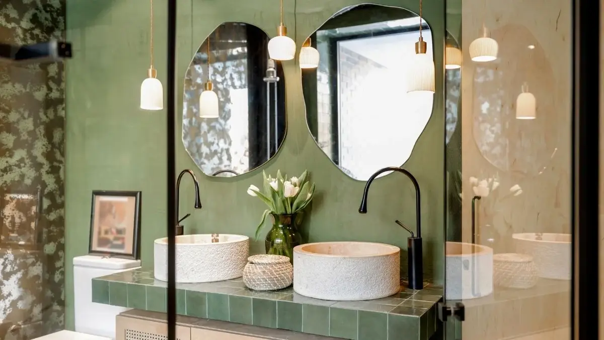 Łazienka z zieloną ścianą, na niej dwa lustra, pod nimi dwie białe umywalki na blacie wyłożonym zielonymi kafelkami