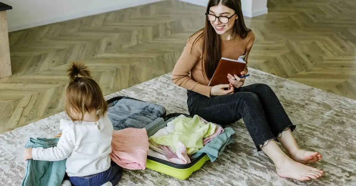 Uśmiechnięta mama z listą rzeczy do spakowania i odwrócona tyłem córeczka siedzą koło spakowanej walizki.