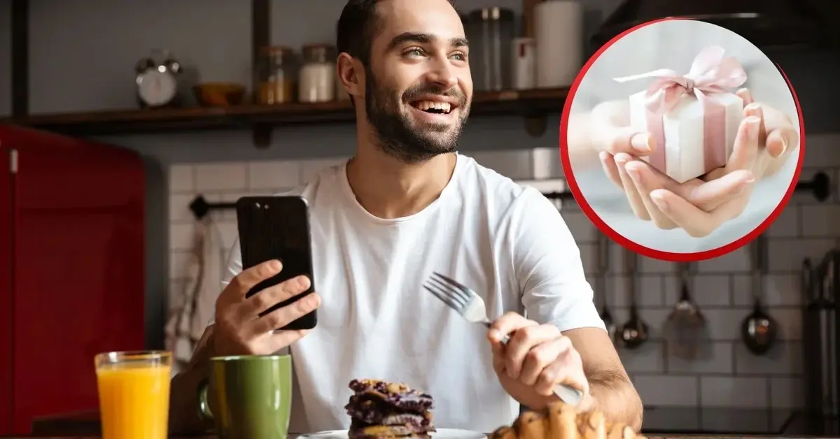 Uśmiechnięty mężczyzna z telefonem je śniadanie. Zdjęcie kobiecych dłoni trzymających prezent na Dzień Mężczyzny.