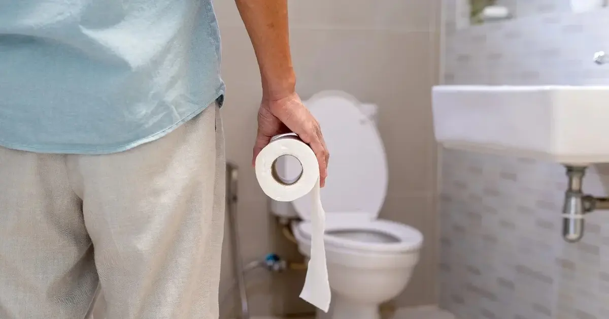 Plecy i nogi mężczyzny idącego do toalety. Mężczyzna trzyma w ręce papier toaletowy.