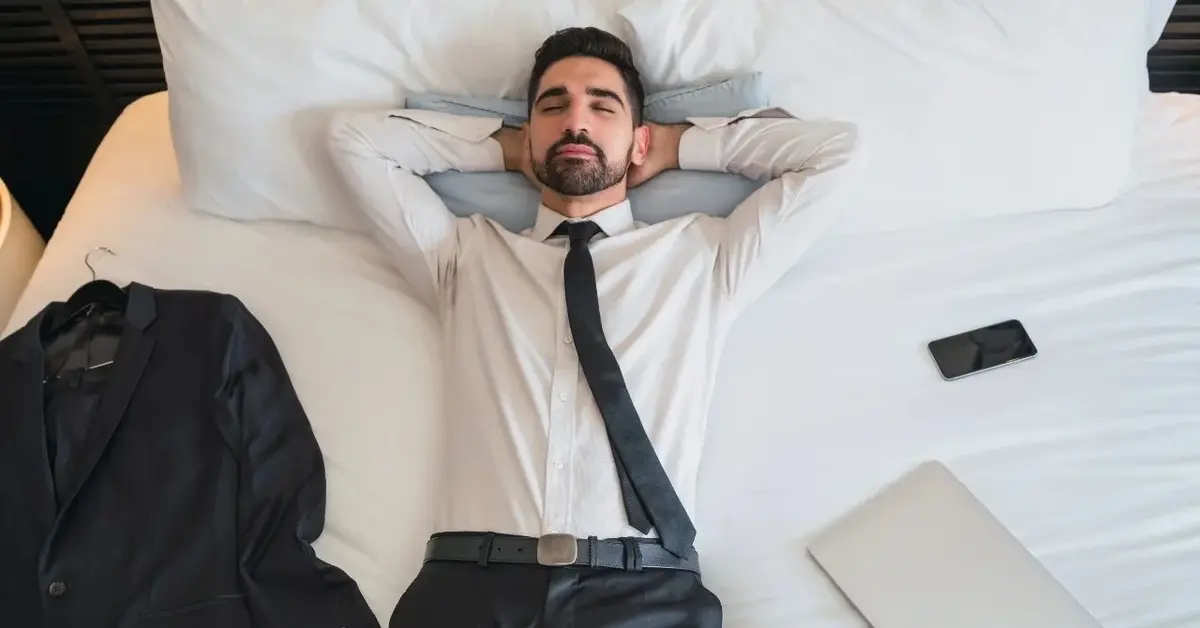 Mężczyzna w formalnym ubraniu leży na łóżku z zamkniętymi oczami i odpoczywa.