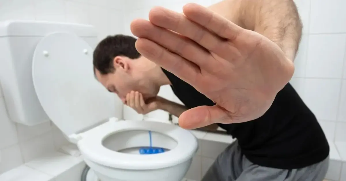 Mężczyzna klęczy przed toaletą i zasłania usta, żeby nie zwymiotować.