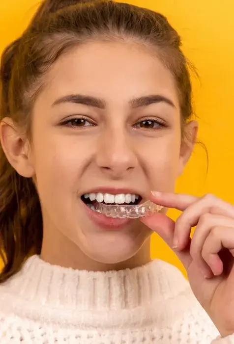 Dziewczynka w białym swetrze z uśmiechem zakłada przeźroczystą nakładkę ortodontyczną