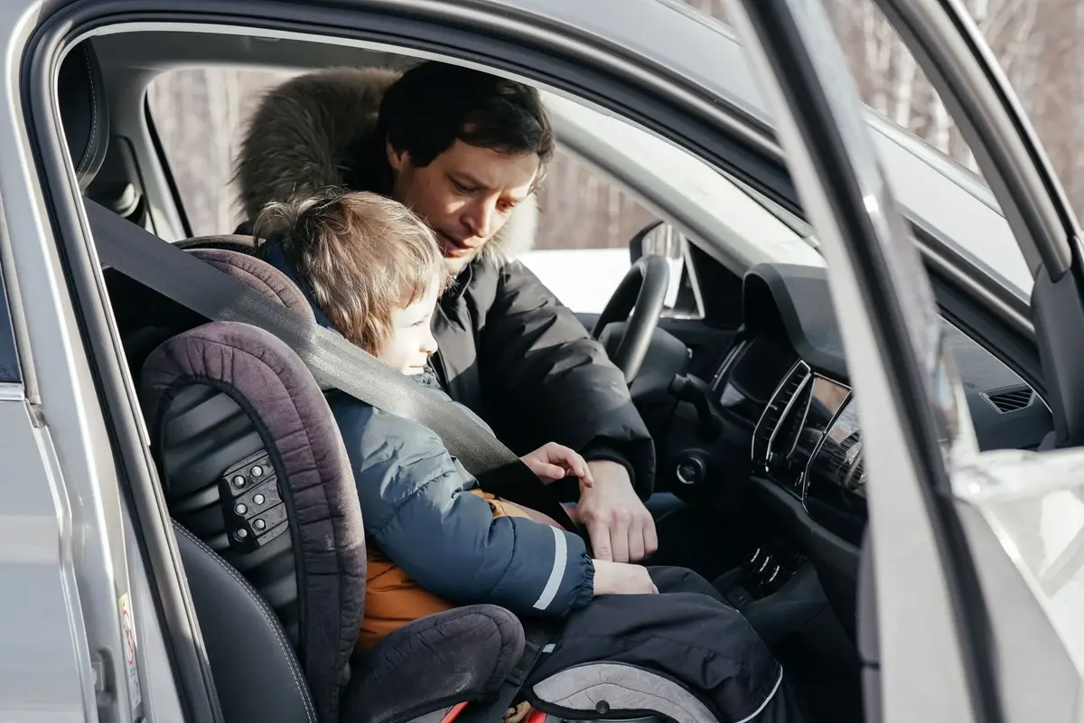 Otwarte drzwi przednie od strony pasażera srebnego samochodu, widać ojca zabezpieczającego dziecko w obrotowym foteliku 