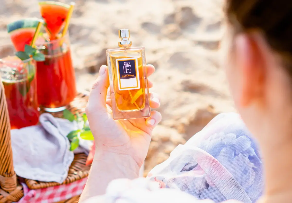 Na plaży siedząc na kocu przy czerwonych drinkach z arbuzem kobieta przygląda się flakonowi odpowiednika perfum