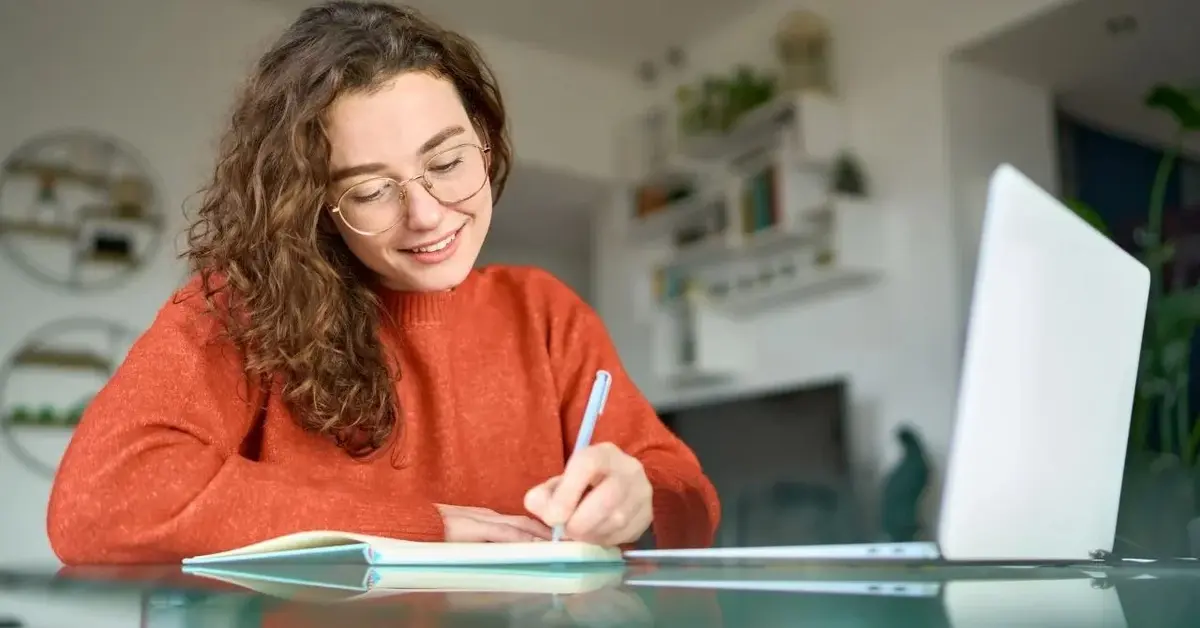 Uśmiechnięta brunetka o kręconych włosach w okularach do czytania notuje w zeszycie przed otwartym laptopem