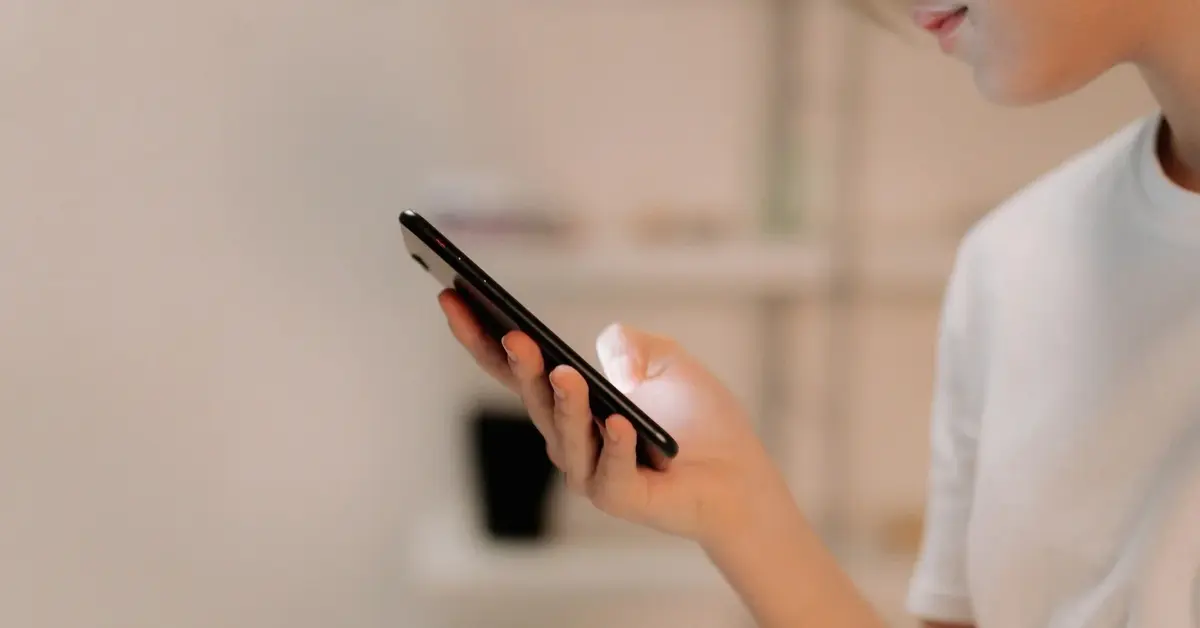 Osoba w białym podkoszulku w domu trzyma w ręku telefon komórkowy