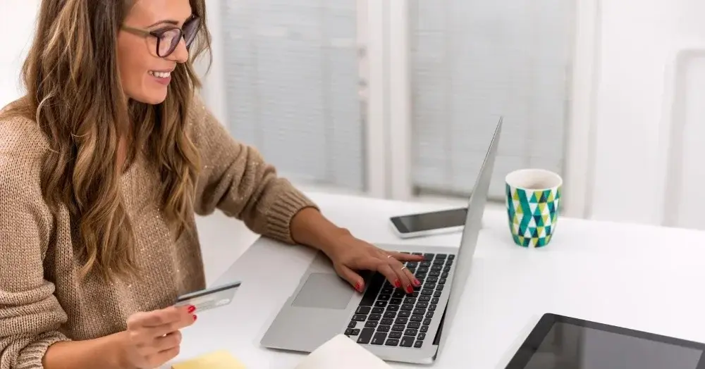 Przy biurku uśmiechnięta kobieta w okularach i beżowym swetrze sprawdza w komputerze jak zadbać o poduszkę finansową