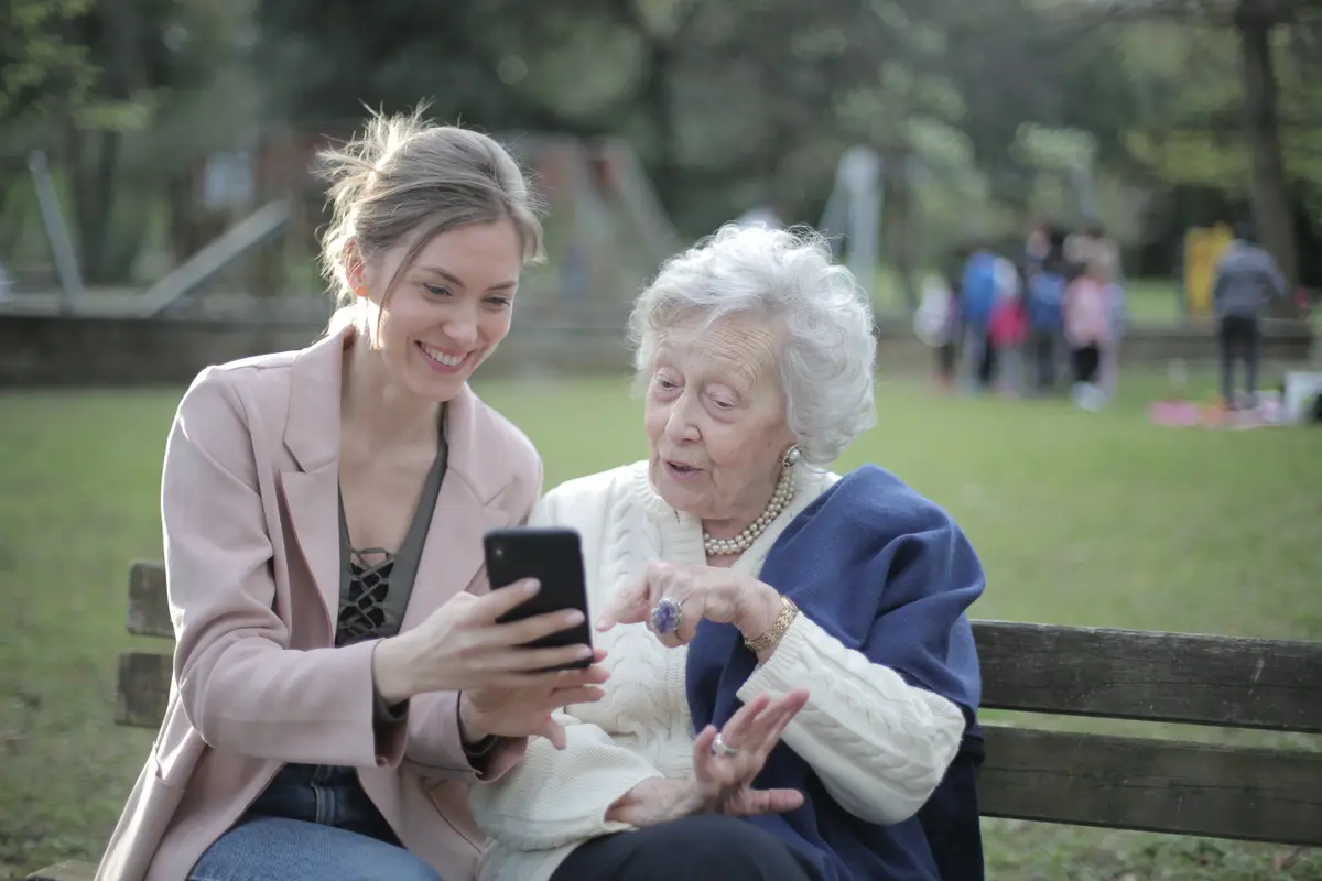 Kobieta opiekunka siedzi na ławce w parku pokazując starszej kobiece ekran smartfona