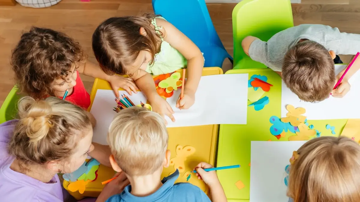 dzieci siedzą przy kolorowych stolikach w przedszkolu i rysują