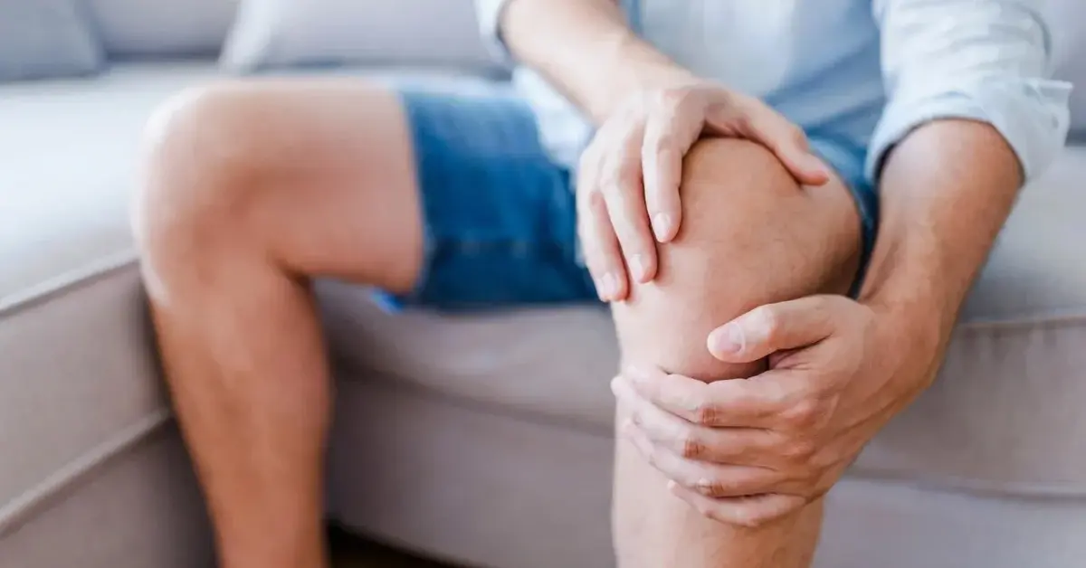 Mężczyzna w krotkich spodniach siedząc na kanapie trzyma się za bolące kolano