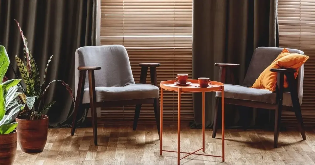 Dwa fotele i pomarańczowy stolik na tle okna z ciemnymi zasłonami i i roletami